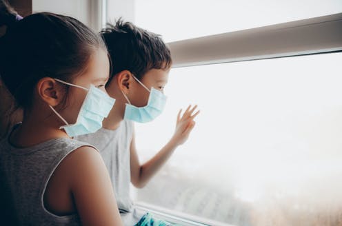 Imagen ilustrativa. Niños aislados usando mascarillas, mirando a través de la ventana.